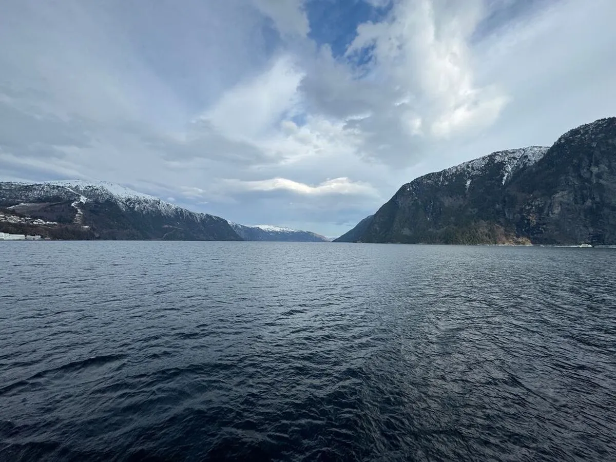 A serene fjord with calm water, surrounded by snow-capped mountains under a partly cloudy sky.