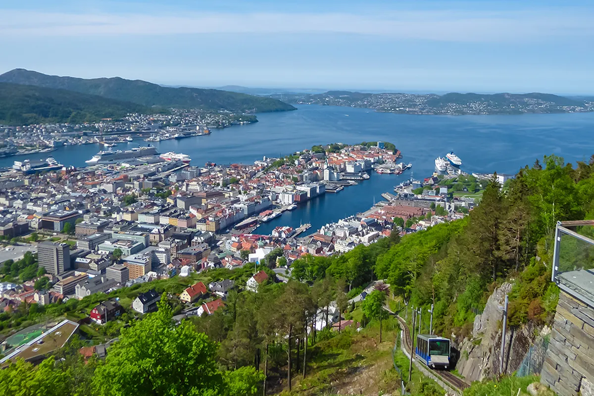 The Fløibanen funicular ascends Mount Fløyen, offering tourists on the viewing platform a spectacular panoramic vista of Bergen, Norway's cityscape, harbor, and fjords on a clear, sunny summer day.