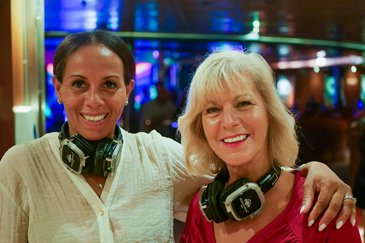 Kerrie Ann and Julie smiling, wearing headphones around their necks, in a vibrant indoor setting with colorful lights in the background.