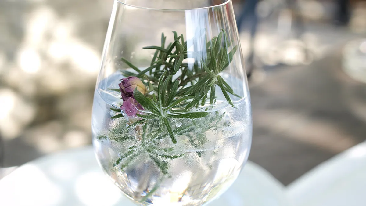 A gin glass garnished with rosemary sprigs and a small purple flower, set on a reflective surface.