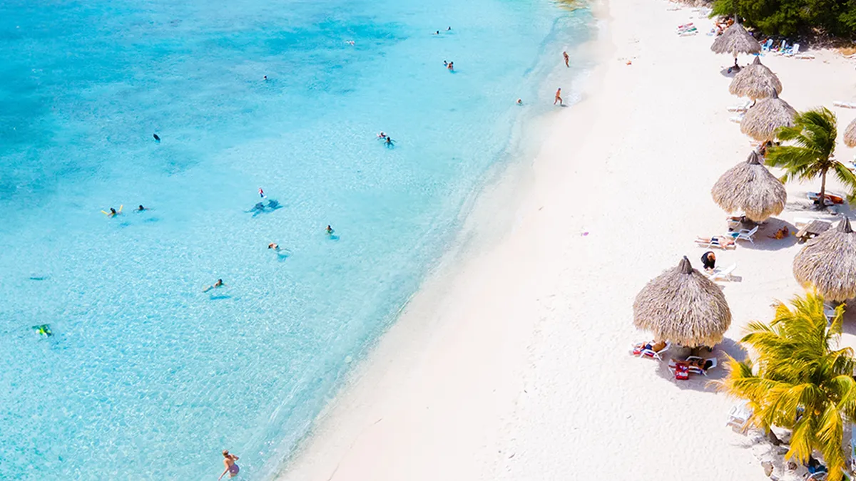 Aerial view of a beach in Curacao with turquoise water, white sand, people swimming, and straw umbrellas surrounded by palm trees.