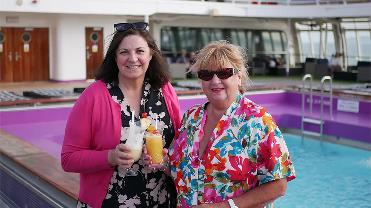 Anne & Yvonne smiling by a pool on the ship, holding tropical drinks.