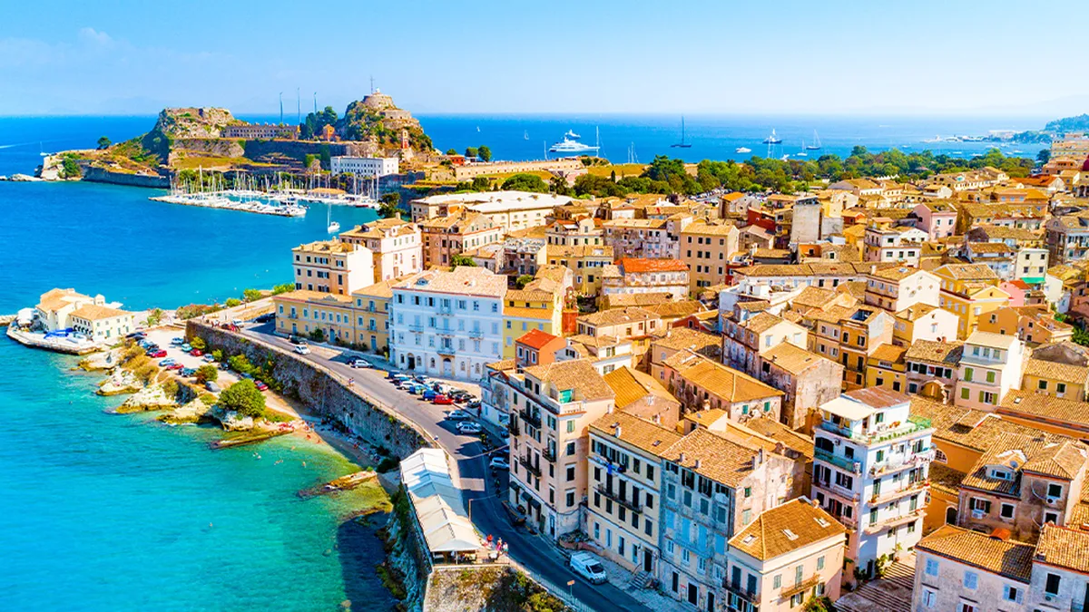 Aerial view of Corfu with colorful buildings, a fort on a hill, and a vibrant blue sea under a clear sky.