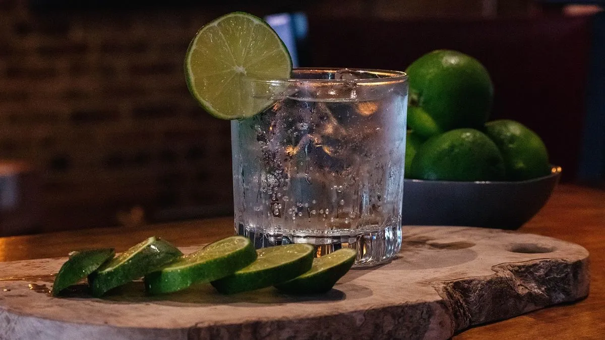 A glass of gin with a lime slice on the rim, surrounded by sliced limes on a wooden board, with whole limes in the background.