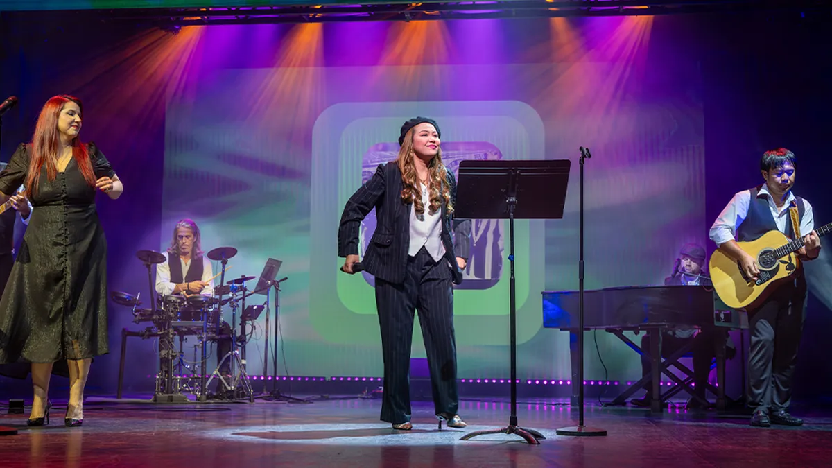 Performers on stage with vibrant lighting; a singer at the center, musicians playing drums, guitar, and keyboard in the background.