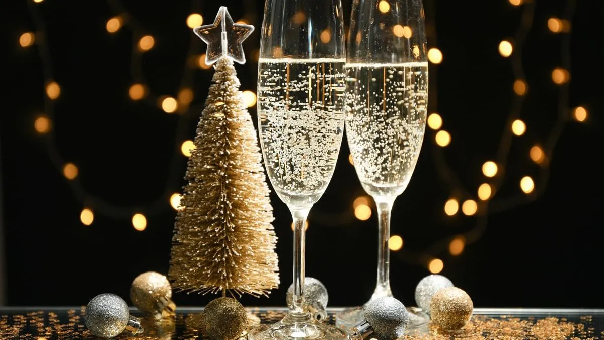 Two champagne glasses with bubbles, a small gold Christmas tree, and festive lights in the background on a reflective surface.