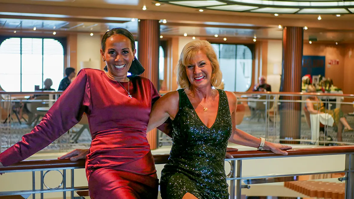Kerrie Ann and Julie in elegant evening dresses pose together, smiling, on a balcony inside a stylish, well-lit venue.