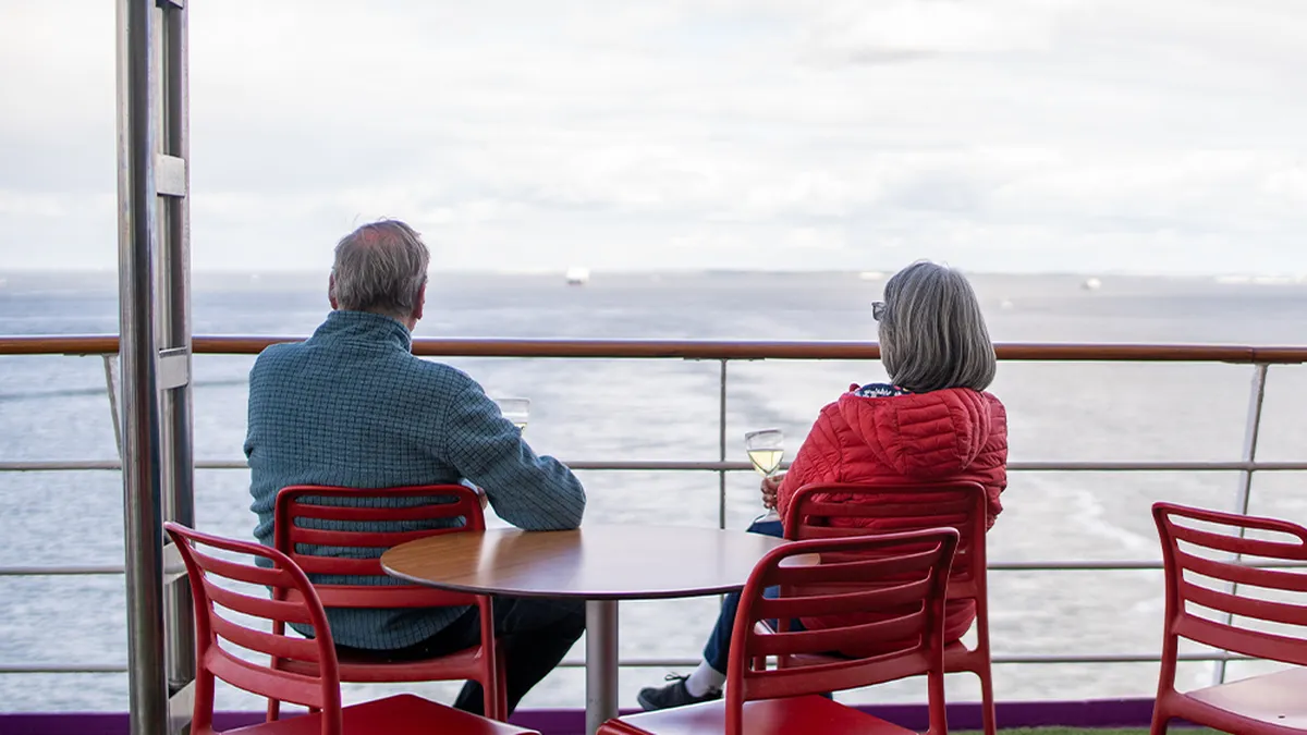 Two people sitting at a table on a ship's deck, looking out at the ocean, holding wine glasses.