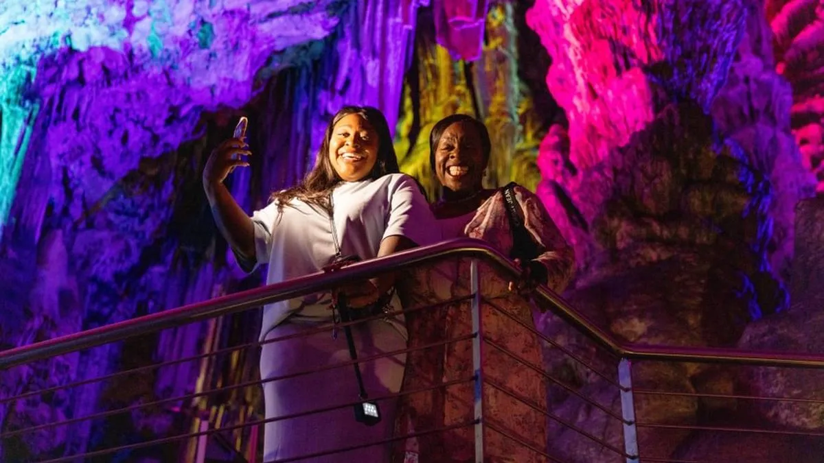 Jacque & Tamara smile on a railing inside a cave, illuminated by colorful lights in pink, purple, and yellow hues.