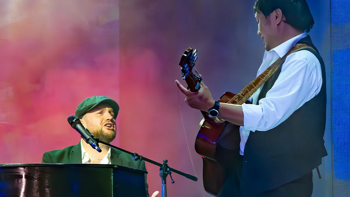 Two musicians perform on stage; one plays the piano and sings, while the other plays an acoustic guitar, set against a colorful backdrop.