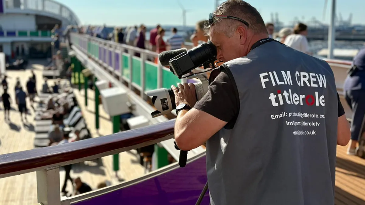Film crew member with a camera filming on a ship's deck, wearing a vest labeled "FILM CREW titlerole." Passengers and deck chairs are visible.
