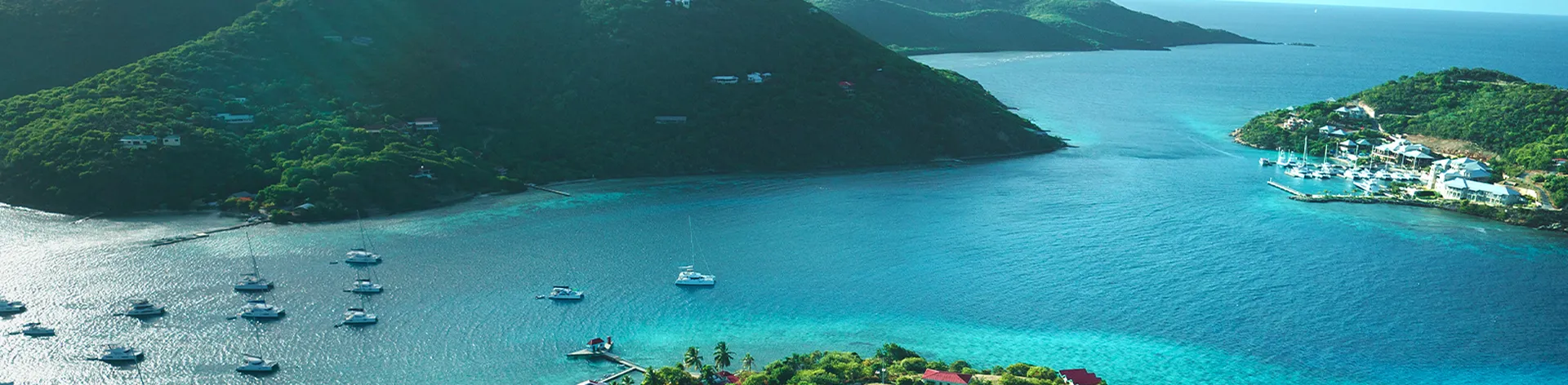 Aerial view of a tropical bay with lush green hills, scattered boats, and vibrant blue water under a clear sky.