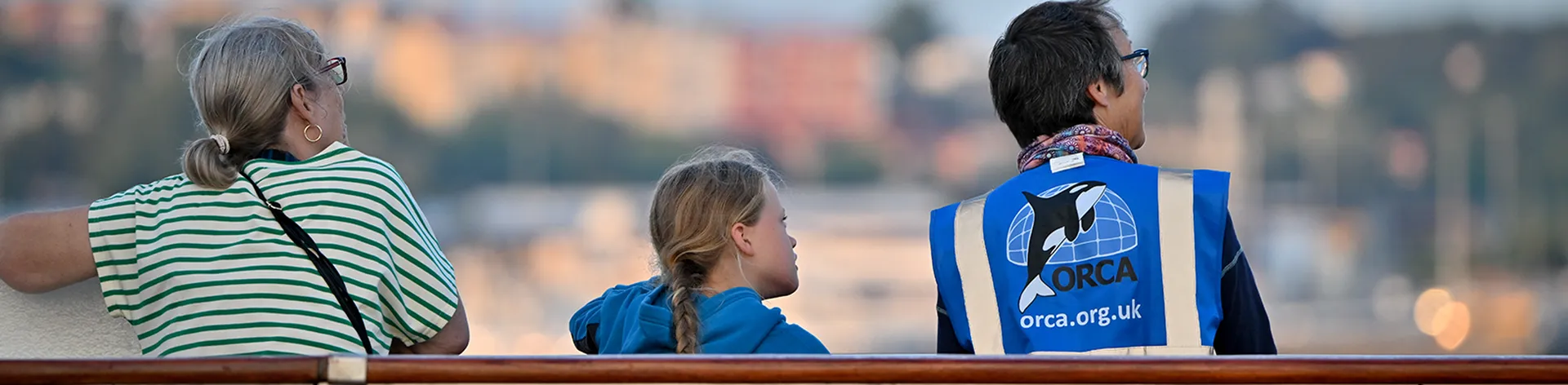 Three people, including a child, stand on a boat deck. One wears an ORCA vest.