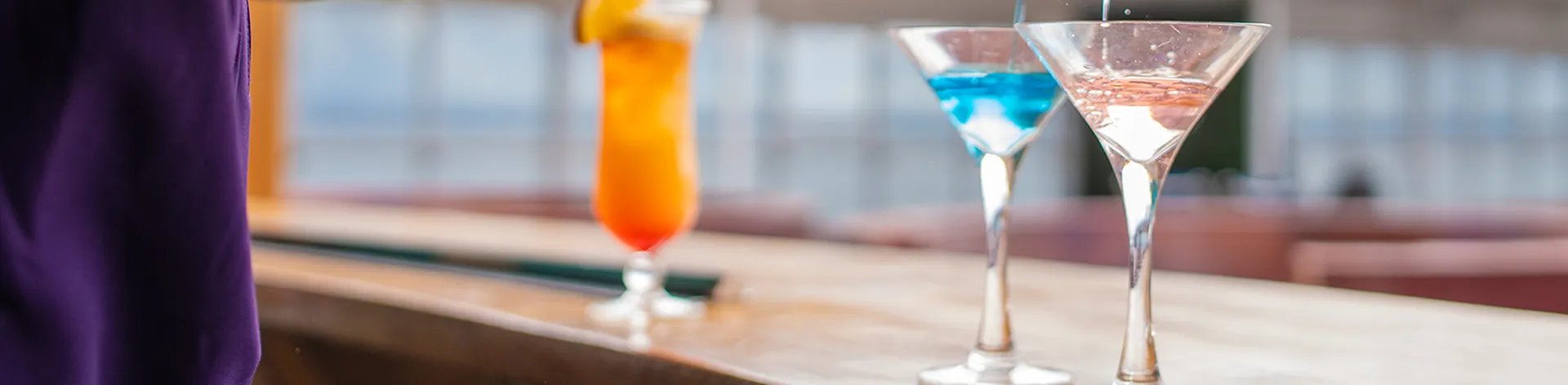 Two martini glasses being filled with blue and pink drinks, with a tall orange cocktail garnished with fruit in the background.