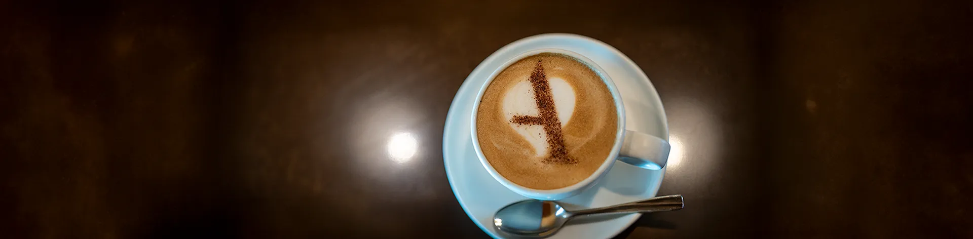 A latte with a letter "A" made of cocoa on the foam, served in a white cup and saucer with a spoon.