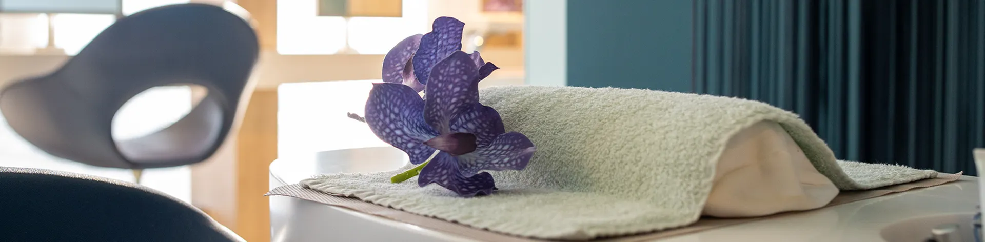 A purple flower rests on a folded towel atop a white surface, with a modern chair and soft lighting in the background.