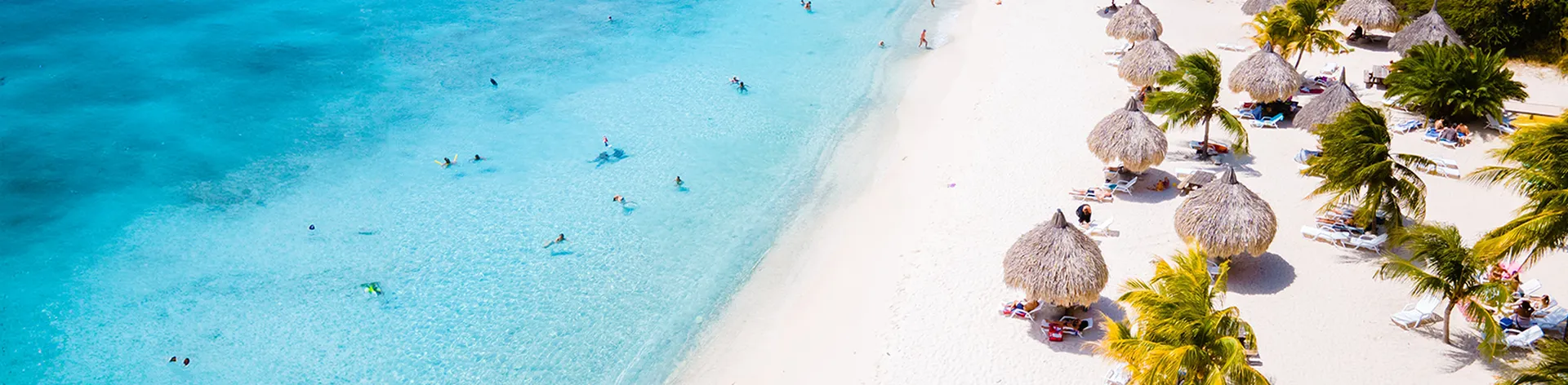Aerial view of a beach in Curacao with turquoise water, white sand, people swimming, and straw umbrellas surrounded by palm trees.