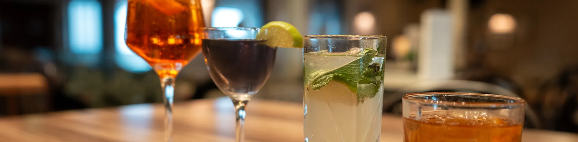 A wooden table displays four cocktails: an orange cocktail, a dark cocktail with lime, a mojito, and a whiskey drink, in a dimly lit room.