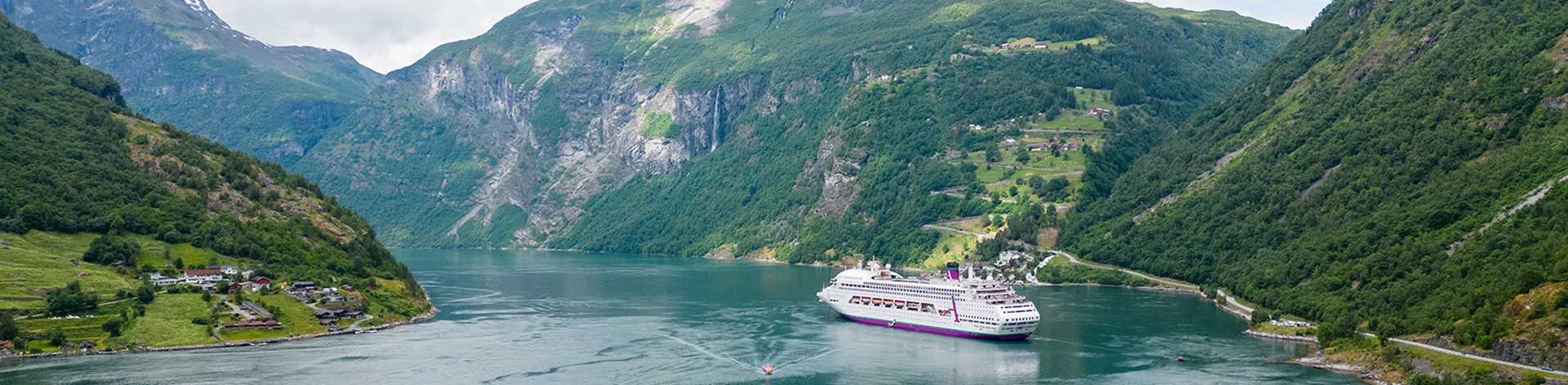 Ambassador Cruise Line ship sailing through a scenic fjord with steep, green mountains and scattered houses