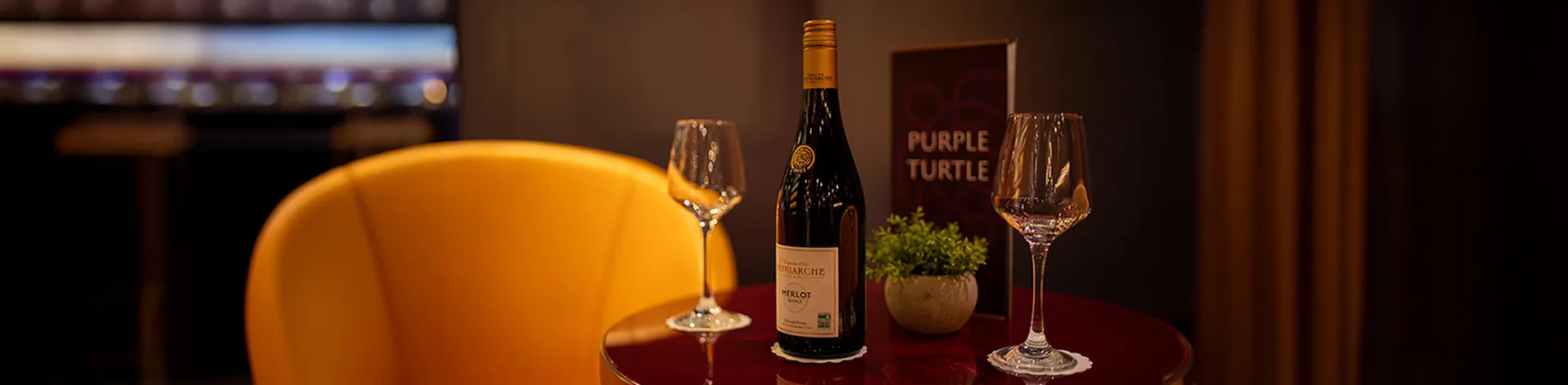 A wine bottle and two glasses sit on a red table, next to a Purple Turtle menu
