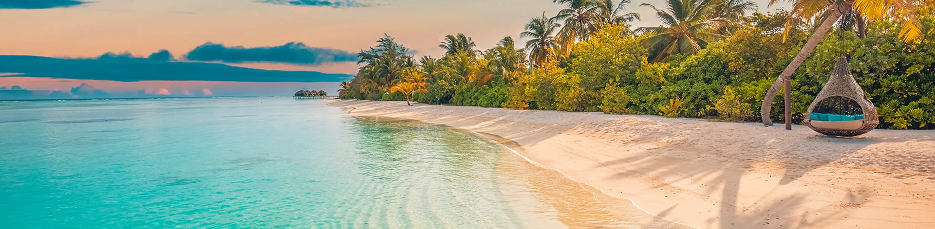 Tropical beach at sunset with calm turquoise water, white sand, lush palm trees, and a hanging wicker chair.