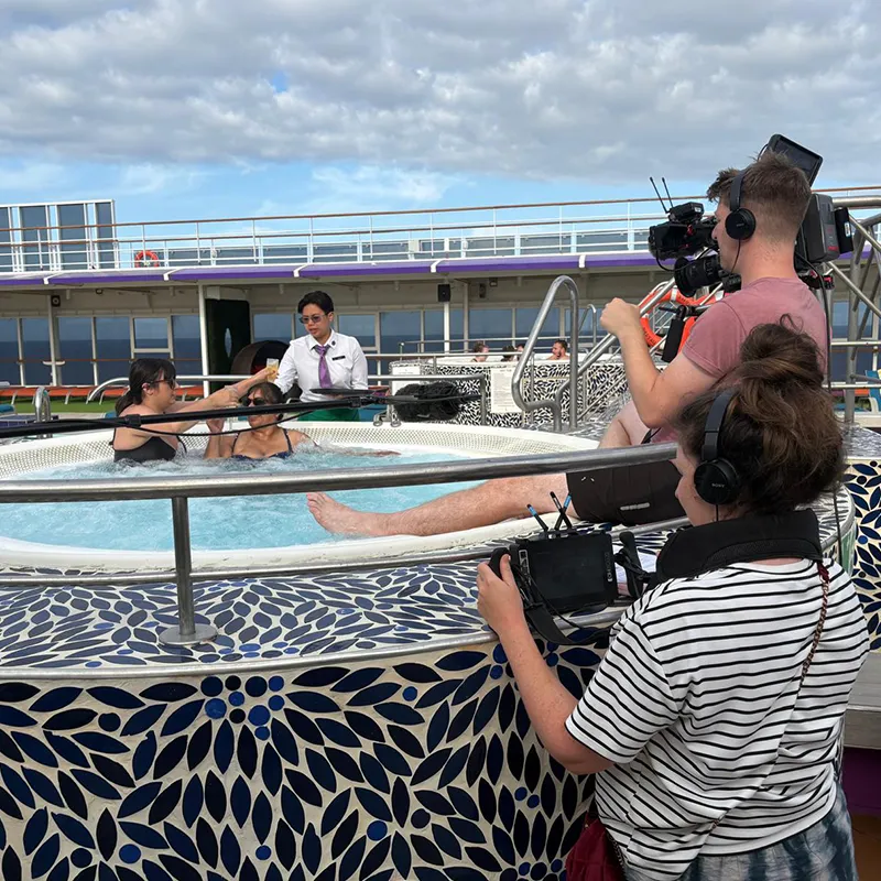 Crew filming people in a hot tub on a cruise ship deck, with a cloudy sky above.