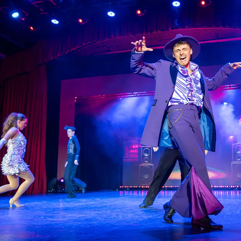 Performer in a vibrant purple suit and hat dances energetically on stage, accompanied by dancers in glittery outfits, under colorful stage lights.
