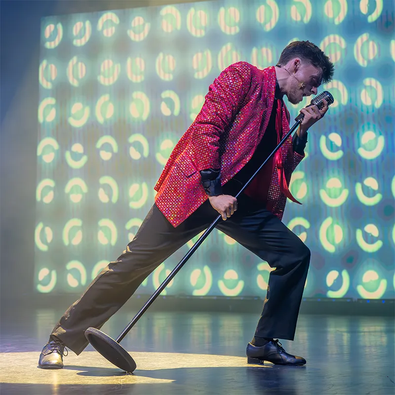 Performer in a red sequined jacket passionately singing into a vintage microphone on stage with a vibrant, patterned background.
