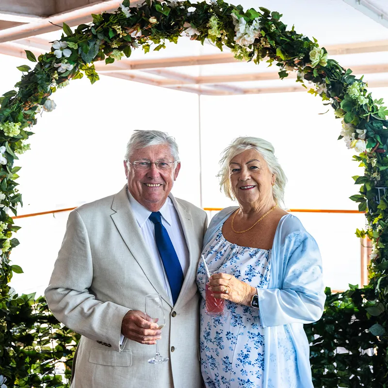 Clive & Bernie smiling under a floral arch, holding drinks.