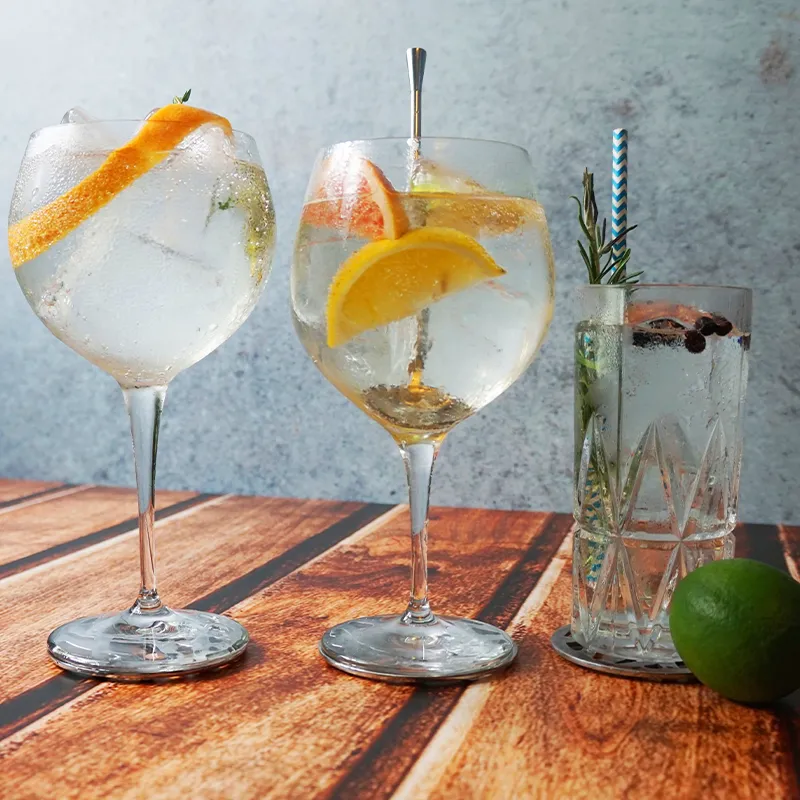 Three cocktails on a wooden surface: two in large glasses with citrus garnishes, and one in a tall glass with herbs and berries, next to a lime.