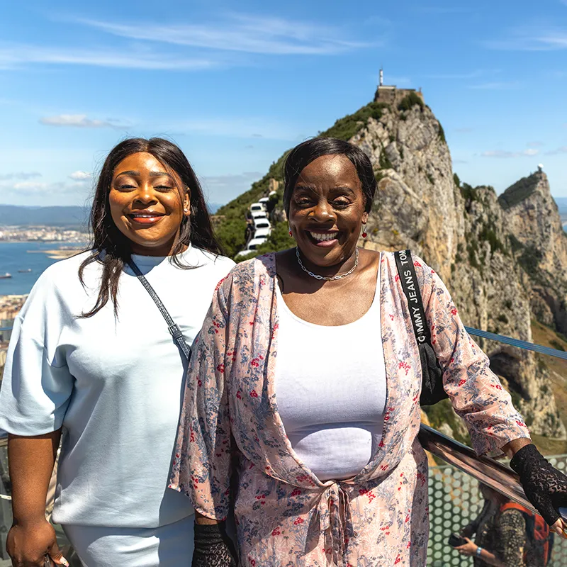 Jacque & Tamara pose on a sunny day with the Rock of Gibraltar in the background, featuring clear skies and a distant view of the sea.