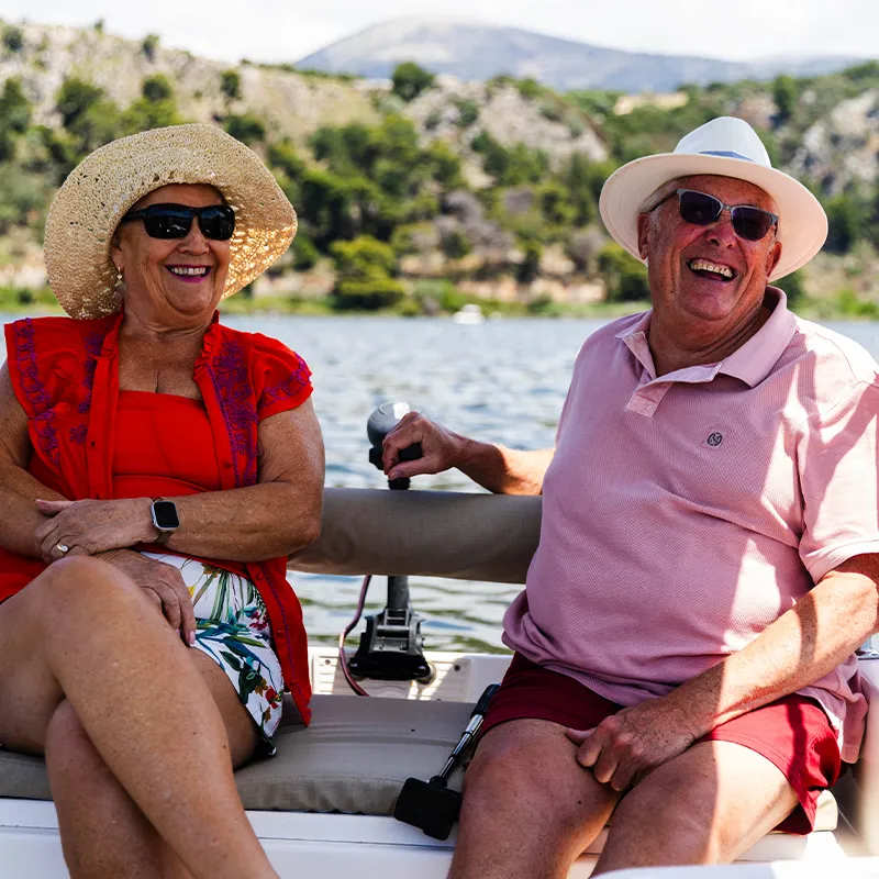 Clive & Bernie smiling on a boat, wearing sun hats and sunglasses, with a scenic lake and mountain backdrop.