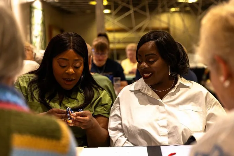 Jacque & Tamara engaged in conversation. One examines a beaded item, while the other smiles. Blurred background with people.