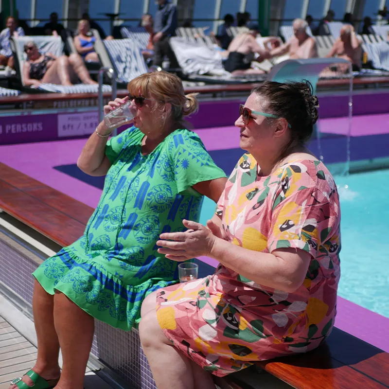 Anne & Yvonne in colorful dresses sit by a pool, one drinking from a glass. Sunbathers relax on lounge chairs in the background.