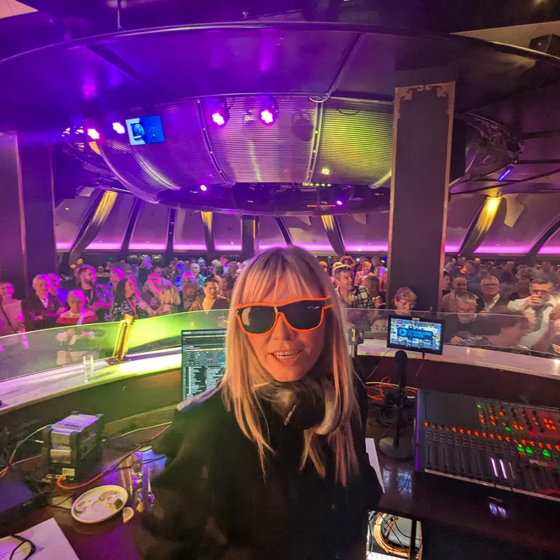 DJ wearing sunglasses performs on stage with a vibrant audience and colorful lighting in the background.