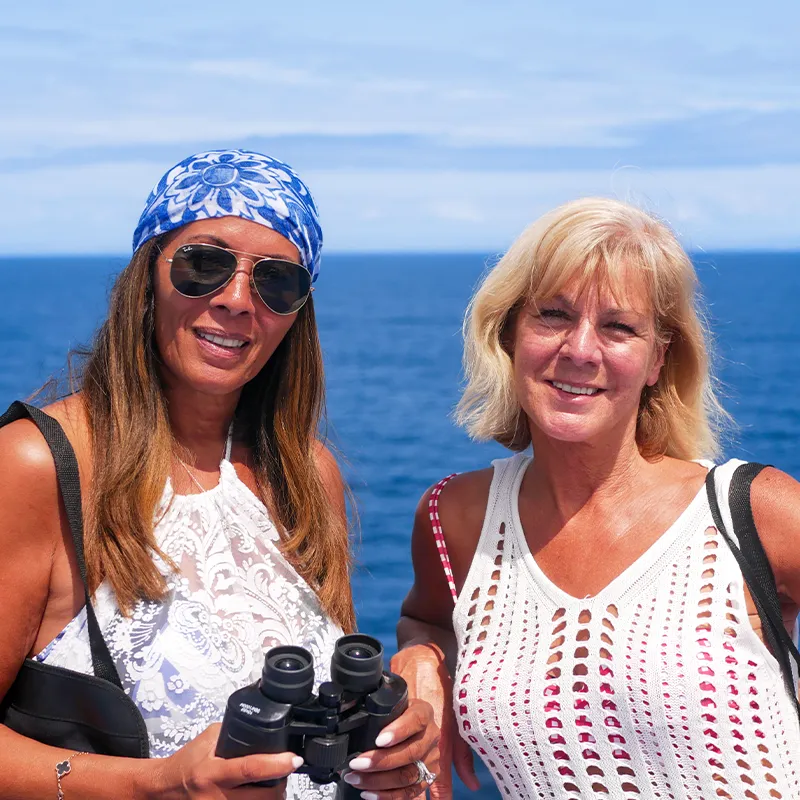 Kerrie Ann and Julie smiling on a boat deck, one holding binoculars. They wear sunglasses and white tops, with the ocean and blue sky in the background.