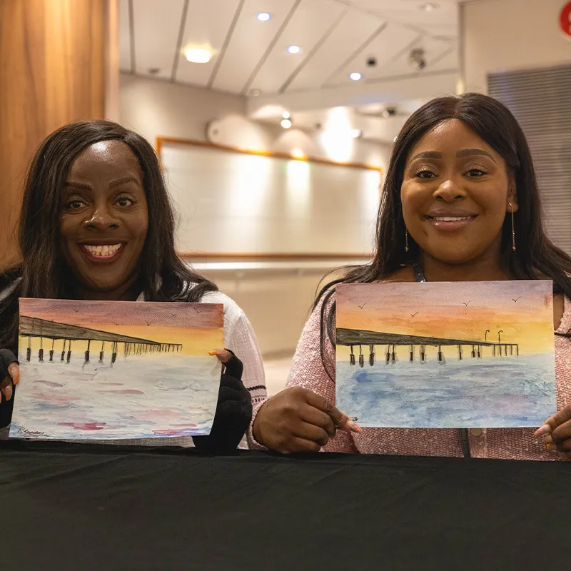 Jacque & Tamara smiling and holding watercolor paintings of a pier at sunset, with birds flying in the sky.