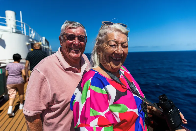 Smiling Clive and Bernie on a ship deck, with the ocean in the background. The woman holds binoculars, and both are enjoying the sunny day.