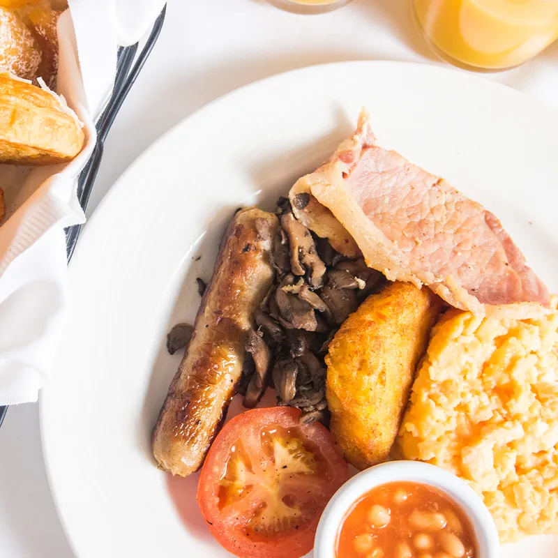 A breakfast plate with bacon, sausage, scrambled eggs, hash brown, mushrooms, tomato, and baked beans. A basket of bread and orange juice are nearby.