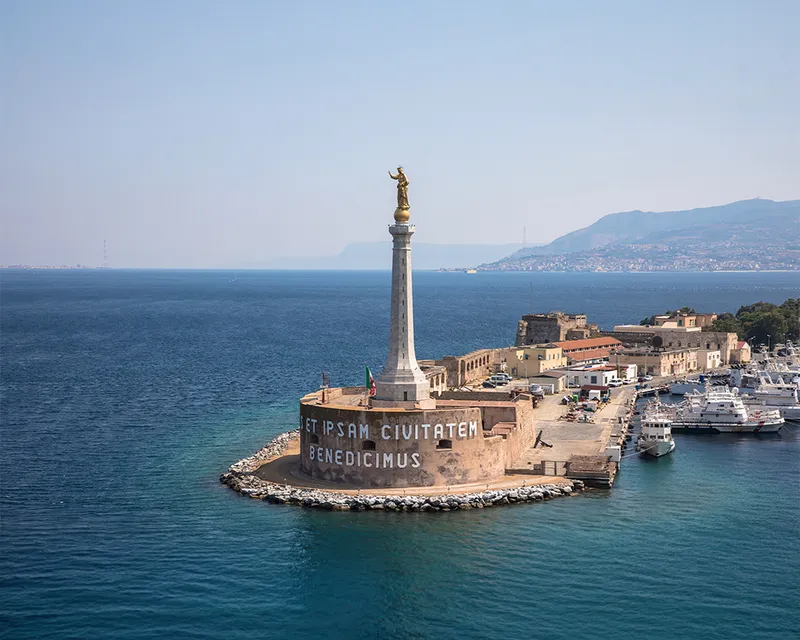 Aerial view of the Madonna della Lettera statue on a tall column at the entrance to Messina's harbor, surrounded by blue sea and distant hills.