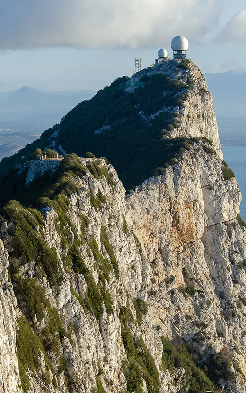 Steep rocky mountain of Gibraltar with radar domes on top, surrounded by lush greenery and a cloudy sky in the background.