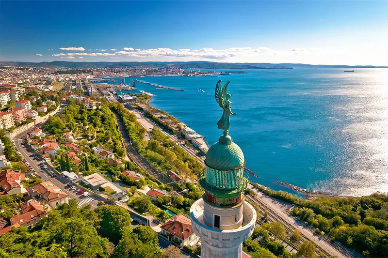Aerial view of Trieste Italy with a prominent statue atop a lighthouse, surrounded by lush greenery, roads, and a vast blue sea under a clear sky.