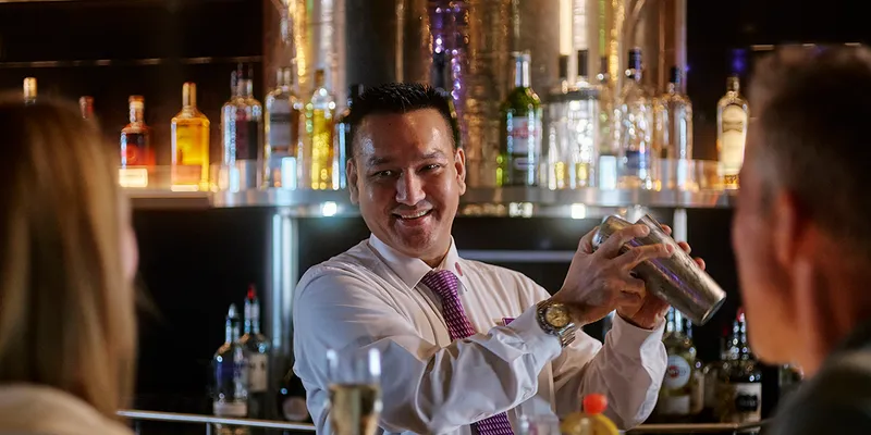 Bartender shaking a cocktail shaker, smiling at guest seated at a bar.