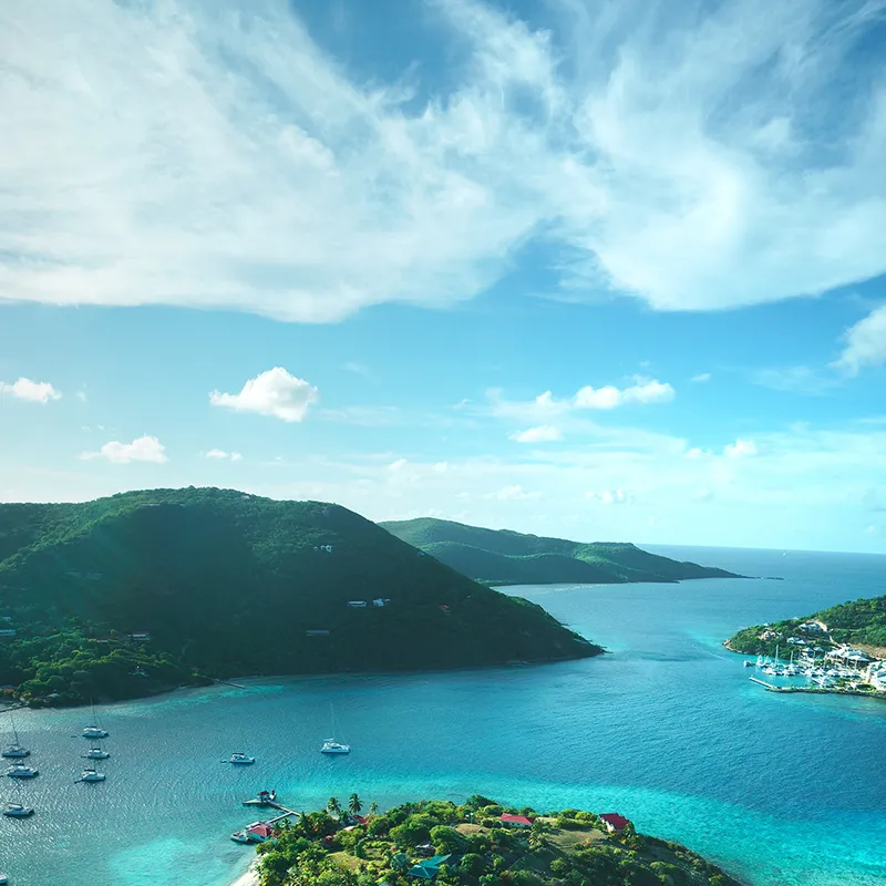 Aerial view of a tropical bay with lush green hills, scattered boats, and vibrant blue water under a clear sky.