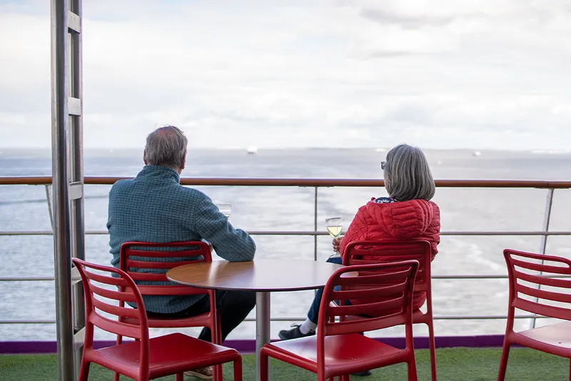 Two people sitting at a table on a ship's deck, looking out at the ocean, holding wine glasses.