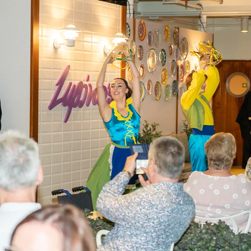 Dancers in colorful costumes perform energetically on Lupino's Stage, with a decorative wall and audience in the foreground.