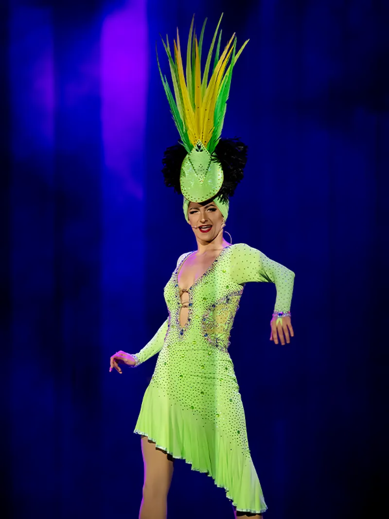 Performer in a sparkling green costume and elaborate headdress dances on stage with a vibrant blue and purple background.