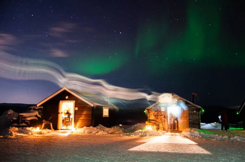 Two wooden cabins in a snowy landscape under a starry sky, illuminated by the green glow of the Northern Lights.