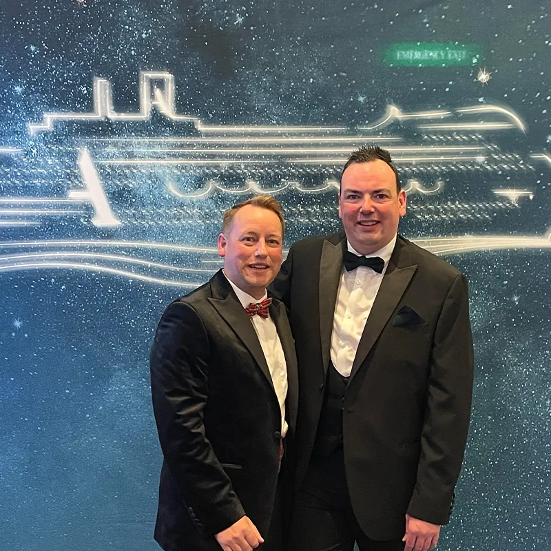 James & Damien in formal suits smiling in front of a starry backdrop with a cruise ship outline.