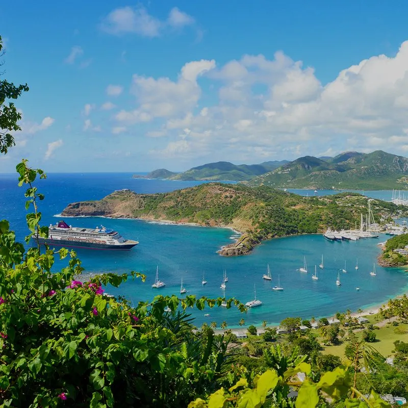 Aerial view of a scenic bay with lush green hills, a large cruise ship, multiple sailboats, and clear blue skies.